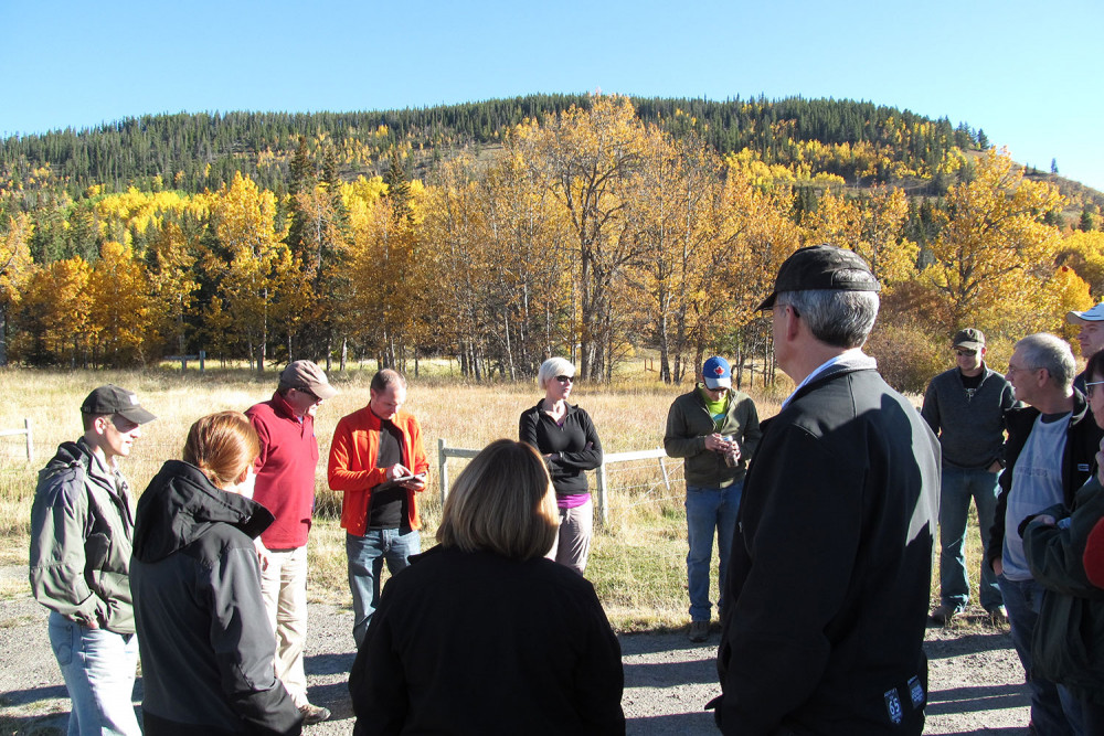 group of people standing on a sunny fall day with a mixed-wood hill in the background